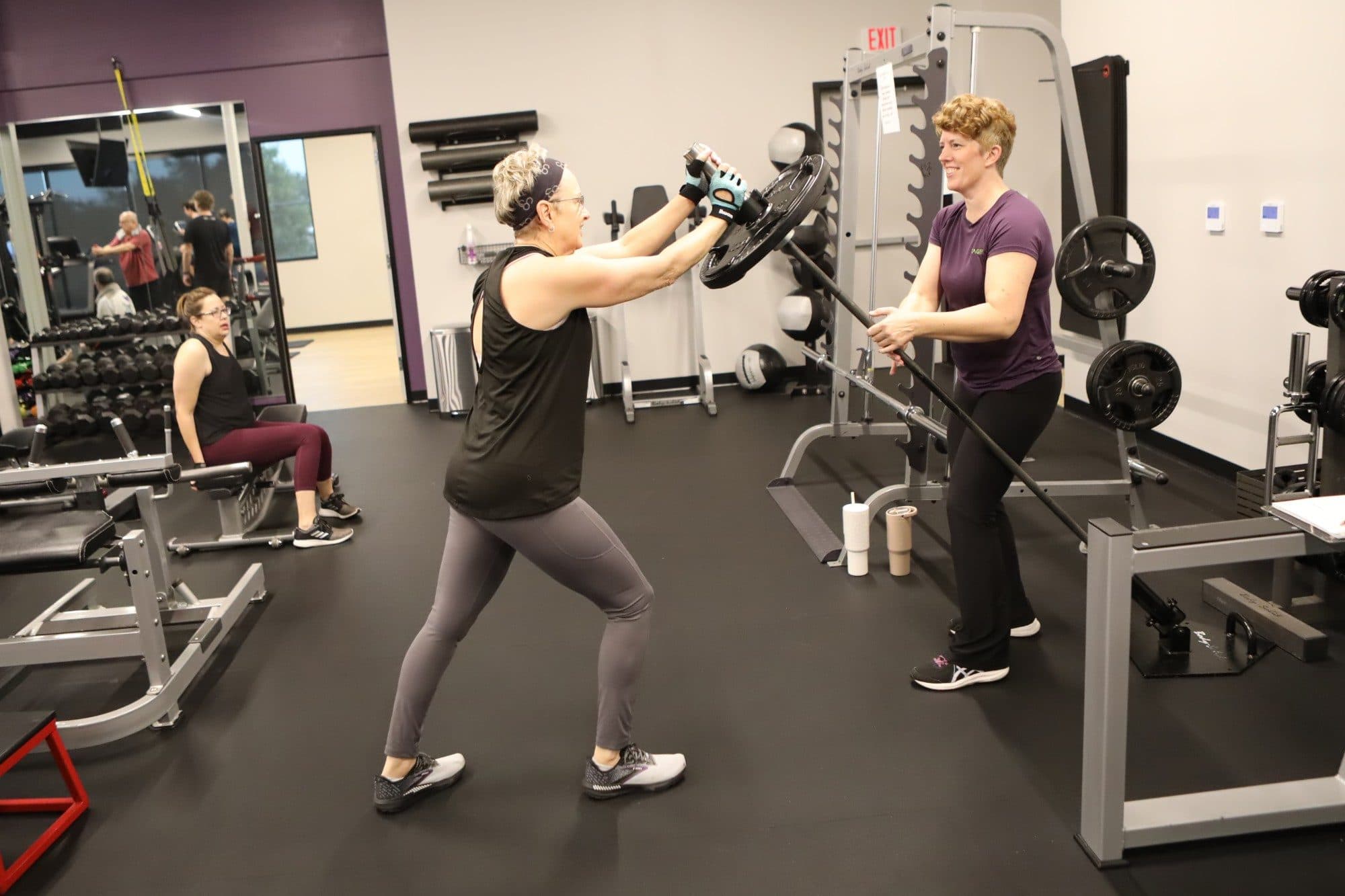 A GetAgeFit client in a lunge pressing a weight plate overhead while her trainer spots her with a barbell at the Georgetown studio